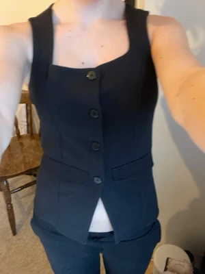 Svart figursydd väst med knappar - Snygg svart väst med fyrkantig urringning och fyra svarta knappar framtill. Figursydd modell med två dekorativa fickor och lätt croppad passform. Perfekt att styla med jeans eller kostymbyxor för en cool look.