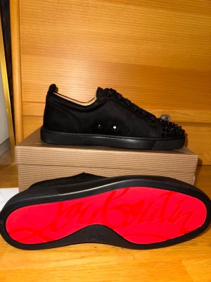 Christian Louboutin svarta sneakers med Spikar  - Svarta sneakers från Christian Louboutin med ikonisk röd sula och spetsig tå. Ovandelen är i mocka med snörning och coola svarta nitar framtill. Insidan är beige och sulan har Louboutin-loggan i rött. Perfekt för dig som gillar edgy designerstil.