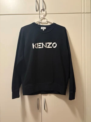 Svart sweatshirt från Kenzo L - Säljer en svart sweatshirt från Kenzo i storlek L. Tröjan har en klassisk passform med rund hals och stort vitt Kenzo-tryck på bröstet. Tillverkad i mjuk bomull och har långa ärmar samt ribbade muddar vid ärmslut och nederkant.