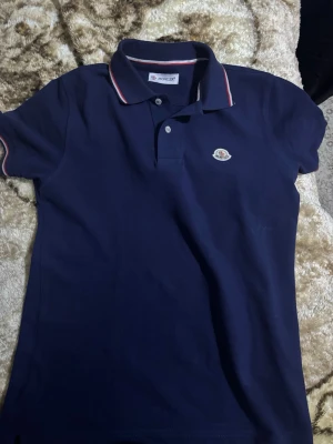 Mörkblå Moncler piké med röd/vit kant - Säljer en mörkblå pikétröja från Moncler med klassisk krage och två knappar framtill. Skulle priset velas diskutera är det bara att skriva till mig men tröjan är också helt oanvänd bara att jag hade tagit bort prislappen innan jag tog bild. 