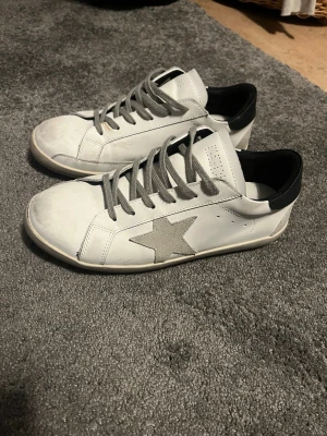 Superstar sneakers med grå stjärna - Snygga vita sneakers från Golden Goose med grå mockadetalj i form av en stjärna på sidan. Skorna har grå snörning, svart häl och platt sula. Insidan är märkt med 'SUPER-STAR'. Perfekt för dig som gillar streetstyle och coola detaljer. Priset kan diskuteras vid snabb affär. Inget og medföljes. Hör av er vid frågor!