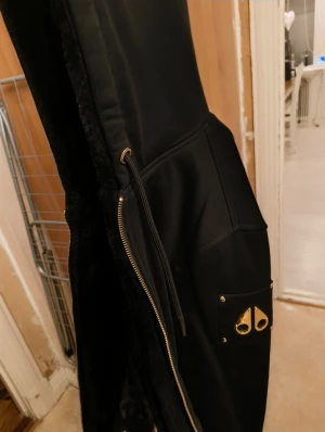 Svart pufferjacka Moose Knuckles - Svart pufferjacka från Moose Knuckles med guldfärgade metalldetaljer på fickan och dragkedja. Jackan har huva, snörning i midjan och quiltad insida med logomönster. Perfekt för dig som gillar streetwear och exklusiva detaljer.