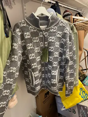 Exklusiv grå stickad kofta från Gucci med vit GG-logga över hela tröjan. Koftan har ribbad hög krage, dragkedja framtill och fickor med vita detaljer. Perfekt för dig som vill ha en lyxig och snygg look med italiensk design. Storlek XL men är liten i storlek så passar og bättre dem som har  L