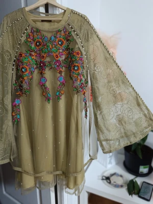 Beige blommig klänning från Zahra Rubab - Superfin beige klänning med färgglad broderad blommig design och pärldetaljer. Vida spetsärmar och transparent mesh ger en lyxig känsla. Perfekt för dig som vill sticka ut med unika detaljer och färger. Märke: Zahra Rubab.