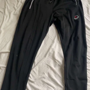 Svarta mjukisbyxor från Moncler - Svarta mjukisbyxor från Moncler med broderad logga på benet och vita snören i midjan. Byxorna har en rak passform och är gjorda i mjukt material som är skönt att ha på sig. Perfekta för chill eller träning.