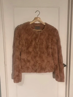 Beige/rosa fuskpälsjacka - Mysig teddyjacka i beige med fluffig yta och rund hals. Jackan har långa ärmar och stängs med silvriga tryckknappar framtill. Insidan är slät för extra komfort. Perfekt för dig som gillar en chill och trendig look.