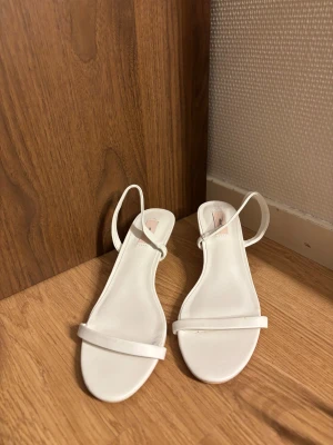 Vita klackar från Nelly - Säljer ett par stilrena vita klackskor från Nelly i storlek 39. Skorna har smala remmar, öppen tå och låg klack som ger en elegant look. Materialet är syntet och designen är minimalistisk med rund tå. Perfekta för dig som gillar clean och modern stil. Aldrig använda!🤍