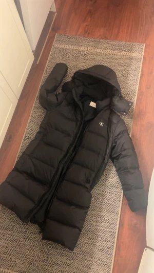 Svart lång pufferjacka Calvin Klein - Säljer en svart lång pufferjacka från Calvin Klein Jeans i storlek XS. Jackan har huva, dragkedja och snyggt CK-logo på bröstet. Perfekt för kalla dagar med sin fluffiga och quiltade design. Materialet är syntet och dun, vilket gör den både varm och lätt.