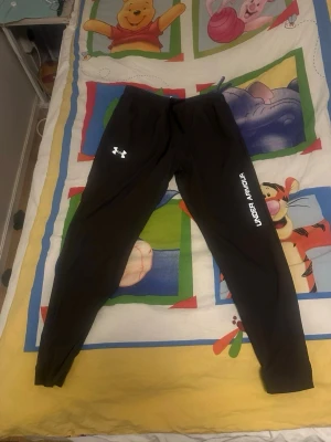 Svarta träningsbyxor från Under Armour - Svarta träningsbyxor från Under Armour med logga på vänster lår och text längs höger ben. Byxorna har snörning i midjan och är gjorda i ett lätt syntetmaterial som passar perfekt för sport och träning. Smal passform och elastisk midja för extra komfort. Strlk XL men passar L
