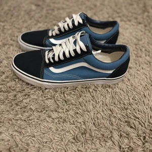 Blå och svarta Vans Old Skool sneakers - Säljer ett par klassiska Vans Old Skool sneakers i blått och svart med vit sula och vit sidosnirk. Skorna har snörning och är tillverkade i canvas och mocka. Perfekta för dig som gillar streetstyle och vill ha en tidlös look. Använda 1 gång 