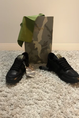 Valentino Garavani svarta sneakers - Svarta sneakers från Valentino Garavani med snygga camouflage-detaljer och platt sula. Skorna har snörning och en robust yttersula med mönstrad struktur. Materialmix av skinn och syntet, perfekt för dig som gillar exklusiv streetstyle.