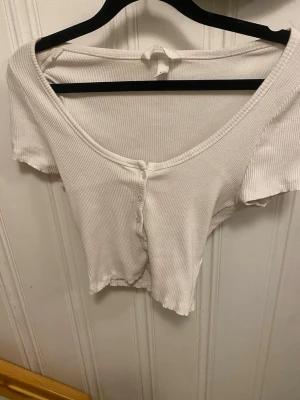 Vit croppad ribbad topp från H&M - Säljer en vit croppad topp från H&M i ribbad bomull. Toppen har korta ärmar, djup v-ringning och knappar framtill. Perfekt för dig som gillar enkel och clean stil med lite edge. Passar dig som vill ha en slim fit och gillar att styla med jeans eller kjol.