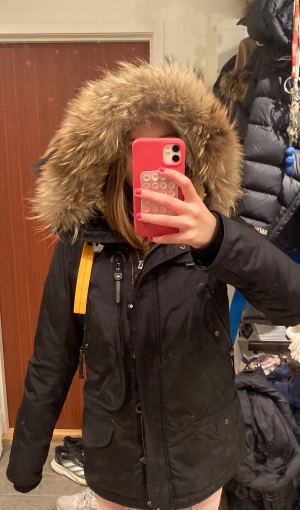 Svart Parajumpers parkajacka med päls - Säljer en svart Parajumpers Anchorage parkajacka i storlek small med stor fluffig päls på huvan och gul detalj vid dragkedjan. Jackan har flera fickor, broderad patch på ärmen och slim fit passform. Perfekt för kalla vinterdagar och riktigt snygg stil. Ord pris cirka 7000 kr