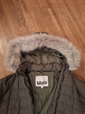 Olivgrön pufferjacka med fuskpäls från Molo - Säljer en olivgrön pufferjacka från Molo med huva och mjuk fuskpäls runt kanten. Jackan har quiltad design, dragkedja framtill och långa ärmar. Perfekt för kalla dagar när du vill hålla dig varm och snygg.