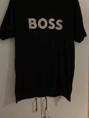Svart BOSS t-shirt med tryck - Svart t-shirt från BOSS med stort vitt BOSS-tryck på bröstet. Klassisk rund halsringning och korta ärmar. Perfekt för dig som gillar stilrena och enkla plagg med tydlig logga. Skön passform och mjukt material.