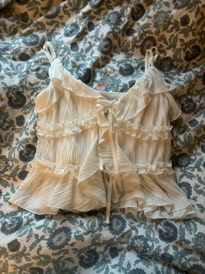 Vit omlott blus med volanger från Amisu - Supersöt vit blus från Amisu i storlek XS. Blusen har tunna axelband, omlott-design med snörning framtill och flera lager volanger. Materialet är lätt och skirt med diskreta ränder, vilket ger en romantisk och feminin vibe. Perfekt för dig som gillar detaljer och en ljus look.