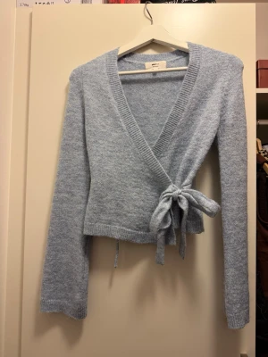 Ljusblå omlottkofta med knyt - Supermjuk ljusblå omlottkofta från Gina Tricot med långa ärmar och bred ribbad kant. Koftan har knytband i midjan som ger en snygg passform och är stickad i ett fluffigt material. Perfekt att slänga på sig när det är lite kyligare ute.