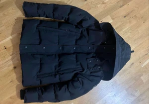 Svart pufferjacka från Canada Goose - Säljer en svart pufferjacka från Canada Goose med huva och snygga detaljer som logopatch på ärmen. Jackan har dragkedja och knappar framtill, samt flera praktiska fickor. Perfekt för kalla dagar och har en clean, modern look.