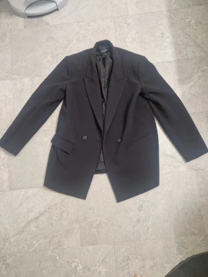 Svart kavaj från KappAhl, storlek 38 - Stilren svart kavaj från KappAhl i klassisk modell med två knappar framtill och snygga fickdetaljer. Kavajen har lång ärm och är fodrad för extra komfort. Perfekt för dig som vill ha en clean och tidlös look. Fåtal gånger använd inga konstigheter.