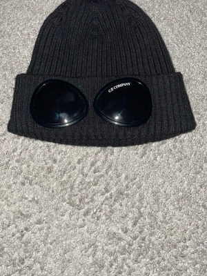 Svart Goggle-mössa från C.P. Company - Svart ribbstickad mössa från C.P. Company med ikoniska goggle-linser på uppviket. Mössan är tillverkad i mjuk ull och har en clean, urban look som sticker ut. Perfekt för dig som vill ha streetstyle med edge och gillar detaljer som syns. Kommer med alla tag och kan scanna