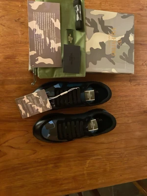 Valentino Garavani camo sneakers - Snygga sneakers från Valentino Garavani med svart och blått camouflage-mönster. Skorna har rund tå, platt sula med mönstrad undersida och svarta snören. Materialet är en mix av skinn och syntet. Kommer med originalkartong, extra snören och dustbag.pris kan diskuteras 