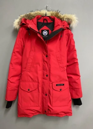 Röd parkas från Canada Goose - Säljer en röd parkas från Canada Goose med huva och snygg pälsdetalj. Jackan har flera fickor med knappar, dragkedja och tryckknappar framtill. Perfekt för kalla dagar med sin långa modell och varma material. 