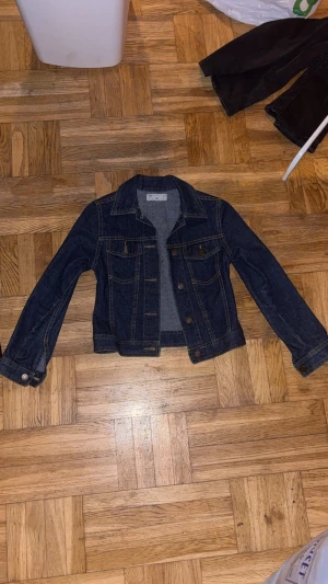 Mörkblå jeansjacka med brunt skinnväst - Säljer en klassisk mörkblå jeansjacka med snygga kontrastsömmar och bruna knappar. Jackan har två bröstfickor och lång ärm. Med följer en cool brun väst i skinn med knappar framtill, perfekt för stil och snygghet