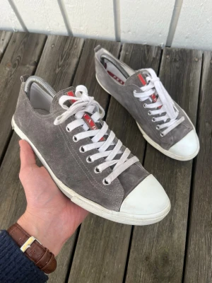 Prada sneakers  - Säljer ett par stilrena grå sneakers från Prada i mjuk mocka. Skick 8/10 riktigt bra, har en liten defekt som ej syns vid användning se bild! Nypris 5000kr men säljs nu för endast 1099kr. Hör av er vid minsta lilla fundering!