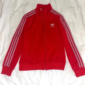Röd Adidas ziptröja med vita ränder - Röd Adidas ziptröja med klassiska vit/lila ränder längs ärmarna och broderad logga på bröstet ❤️Tröjan har hög krage, dragkedja hela vägen och två fickor med dragkedja. Perfekt för en häftig och avslappnad stil 🤍