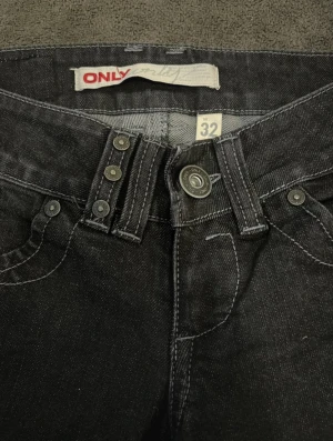 Svarta jeansbyxor från ONLY, W32 L34 - Snygga svarta jeans från ONLY med raka ben och klassisk femficksdesign. Byxorna har låg midja, silverfärgade knappar och detaljerade sömmar. Materialet är slitstarkt jeans och passformen är normal. Perfekt för dig som gillar stilrena och tidlösa jeans.