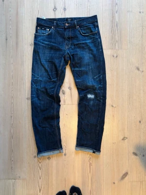 Mörkblå jeans från Hugo Boss 33/32 - Snygga mörkblå jeans från Hugo Boss i klassisk straight fit. Jeansen har coola slitningar och en patchad detalj på ena benet. Femficksmodell med bruna sömmar och läderpatch bak i midjan. Perfekt för dig som gillar en stilren men edgy look.    Längd 101 midja 42 benöppning 20