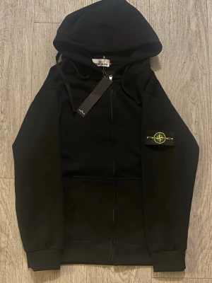 Stone island zip svart - Hej, säljer denna fina svarta zip hoodien från Stone island i storlek M, med inga defekter även helt nytt med prislapp! Hör av er om ni är intresserade!☺️