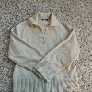 Beige stickad polotröja Gina Tricot - Snygg beige stickad polotröja från Gina Tricot i storlek M. Tröjan har en bred krage, v-ringad öppning och långa ärmar med ribbade muddar och nederkant. Perfekt för att ge din outfit en chill och clean vibe.