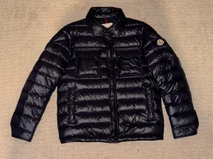 Svart Moncler dunjacka med fickor - Snygg svart dunjacka från Moncler med glansig finish, klassisk krage och flera praktiska fickor framtill. Jackan har både dragkedja och knappar, samt Moncler-logga på ärmen. Perfekt för kalla dagar och ger en clean, modern look.