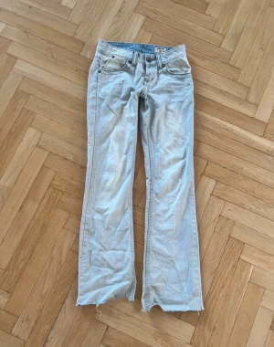 Ljusblå bootcut jeans med rå kant - Säljer ett par ljusblå jeans med bootcut passform och rå, fransig kant nedtill. Klassisk femficksmodell med knappgylf och bälteshällor. Jeansen har en avslappnad vibe och är tillverkade i mjukt denimtyg. Perfekta för dig som gillar vintagekänsla och en chill look.