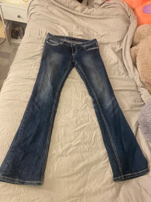 Blå bootcut jeans med broderi från Premiere - Snygga blå jeans från Premiere med bootcut passform och låg midja. Jeansen har detaljerade broderier och paljetter på fickorna både fram och bak, samt kontrastsömmar. Materialet är klassisk jeans med stretch och silverfärgade knappar.