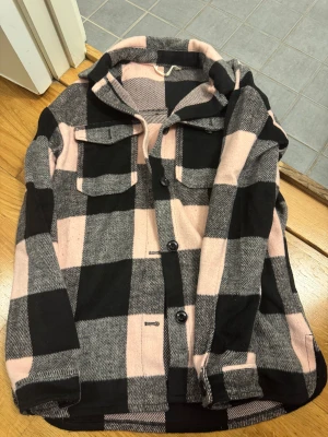 Rutig jacka  i rosa och svart - Säljer en snygg jacka med stora rutor i rosa, svart och grått. Jackan har klassisk krage, två bröstfickor med lock och knappar framtill. Perfekt att slänga över en hoodie för en chill look.