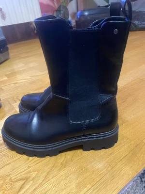 Svarta chunky boots i skinn - Säljer ett par svarta boots med chunky sula och rund tå. Skorna är i skinn med elastiska paneler på sidorna för enkel påtagning. Perfekta för dig som gillar en tuff och stilren look. Passar till både jeans och kjol för en edgy vibe.