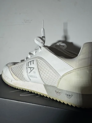 Vita sneakers från Emporio Armani EA7 - Snygga vita sneakers från Emporio Armani EA7 med meshpaneler på sidorna, logotyp på sidan och bak, samt detaljer i syntet och skinn. Skorna har snörning och en platt sula med räfflad undersida för extra grepp. Perfekt för dig som gillar stilrena och sportiga skor.