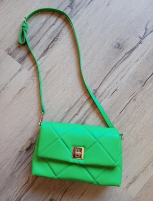 Grön quiltad axelväska från H&M - Snygg axelväska i starkt grön färg från H&M med quiltad design och guldfärgat lås framtill. Väskan har en lång justerbar rem och är tillverkad i syntetmaterial. Perfekt för dig som vill sticka ut med en cool accessoar.