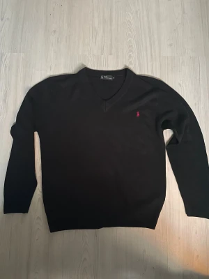 Svart stickad tröja Polo Ralph Lauren - Svart stickad tröja från Polo Ralph Lauren med klassisk v-ringning och röd broderad logga på bröstet. Långärmad och i mjukt material som passar perfekt till kyliga dagar. Enkel och stilren design som funkar till många olika looks.