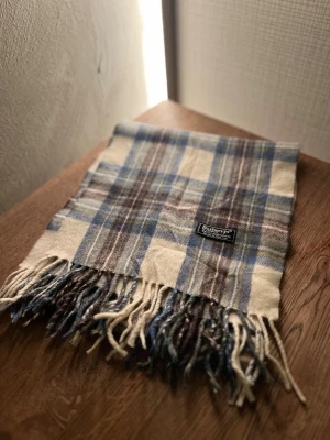 Rutig ullhalsduk från Burberrys - Klassisk rutig halsduk från Burberrys i 100% ull. Halsduken har fransar i ändarna och den ikoniska Burberrys-etiketten. Perfekt accessoar för att lyfta din outfit. Anledningen att de stör ”burberrys” är för att de är en äldre modell. FRI FRAKT BARA IDAG 