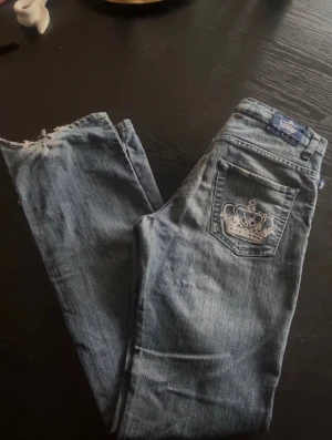 Victoria Beckham blå jeans med broderi - Säljer dessa snygga blå jeans från Victoria Beckham med raka ben och broderade kronor på bakfickorna. Klassisk femficksmodell och midja med knapp. Perfekt för dig som gillar detaljer och streetstil. Se sista bilden för slitskador!! Säljer för att de ej kommer till användning