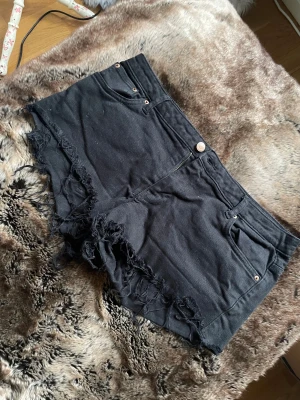 Svarta jeansshorts med fransad kant - Svarta jeansshorts  med fransad och rå kant. Klassisk femficksmodell med knapp och dragkedja framtill. Shortsen har bälteshällor och en slim passform som sitter snyggt på kroppen. Perfekta för varma dagar och en chill streetstyle.