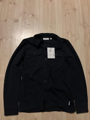 Blå overshirt från Only & Sons - Stilren blå overshirt från Only & Sons med klassisk krage och två bröstfickor med knappar. Skjortan har lång ärm och knäppning framtill, perfekt för lager-på-lager. Materialet är mjukt och känns skönt mot huden. Snygg och enkel att matcha med dina favoritjeans. Aldrig använd