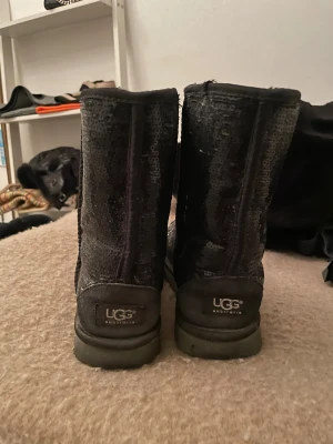 UGG Svarta glittriga - Säljer ett par svarta UGG boots med glittriga paljetter över hela skon. De har en rund tå och platt sula, samt UGG-logga bak på hälen. Insidan är mjuk och varm, perfekt för kalla dagar. Snygga att styla till jeans eller leggings.