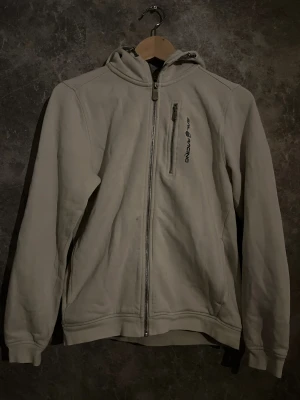 Vit hoodie från Sail Racing med dragkedja - Vit hoodie från Sail Racing med hel dragkedja framtill och en extra zipficka på bröstet. Tröjan har broderad logga på både bröstet och huvan, samt snörning i huvan. Mjuk insida och ribbade muddar vid ärmslut och nederkant. Perfekt för chill dagar.