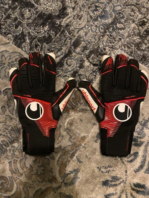 Uhlsport svarta och röda målvaktshandskar - Säljer ett par målvaktshandskar från Uhlsport i svart med röda och vita detaljer då jag inte använder dem längre. Inte använd så mycket, lite färg på högra handflatan men det går att tvätta bort. Köpt för 800 ish, priset kan diskuteras, skriv om ni har frågor 