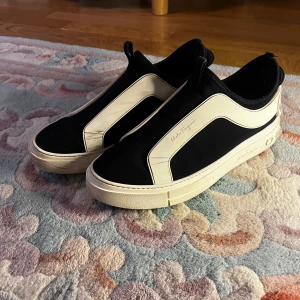 Svarta och vita sneakers från Salvatore Ferragamo - Säljer ett par svarta och vita sneakers från Salvatore Ferragamo med tydliga logodetaljer på hälen och sidan. Skorna har en stilren design med elastiska partier och tjock vit sula. Perfekta för dig som gillar exklusiva och trendiga sneakers. de är storlek m vet inte riktigt vad det är för storlek i eu men jag har storlek 41 och de passar 