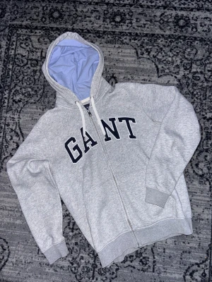 Grå zip hoodie GANT - Säljer en grå hoodie från GANT i storlek M. Tröjan har dragkedja, huva med ljusblå insida och vita snören. Stora GANT-bokstäver i mörkblått framtill. Perfekt för en chill och sportig stil.